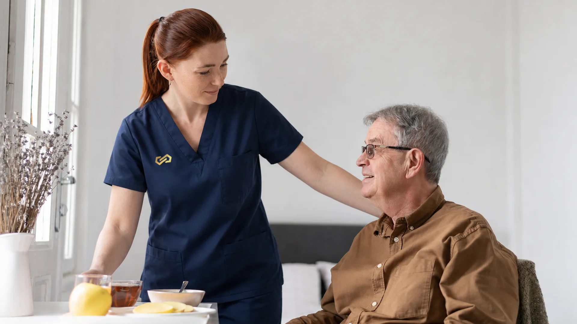 A nurse rests a hand on an older man's shoulder as she sets down his breakfast