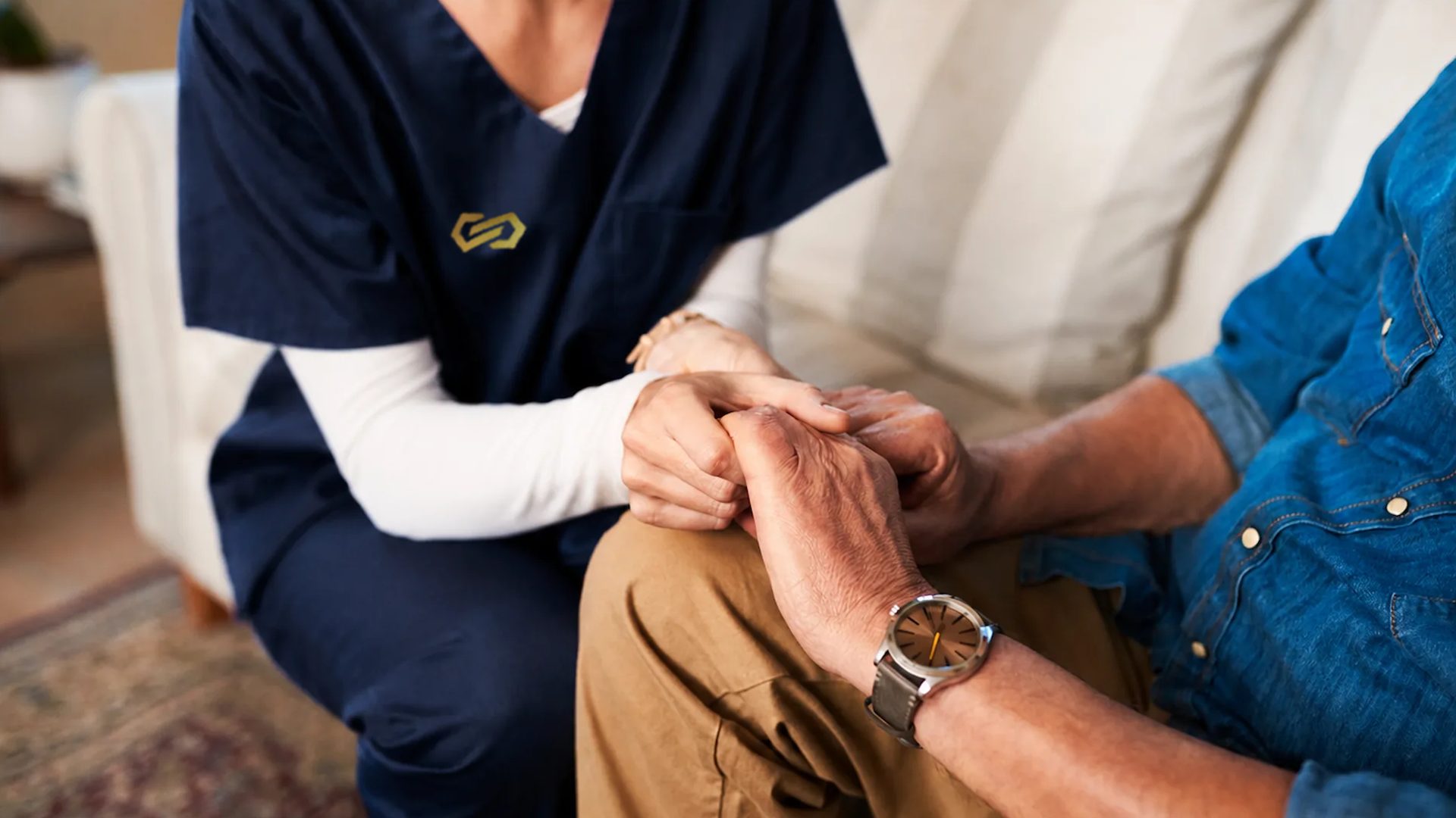 A nurse holds an older man's hands in hers as they sit together