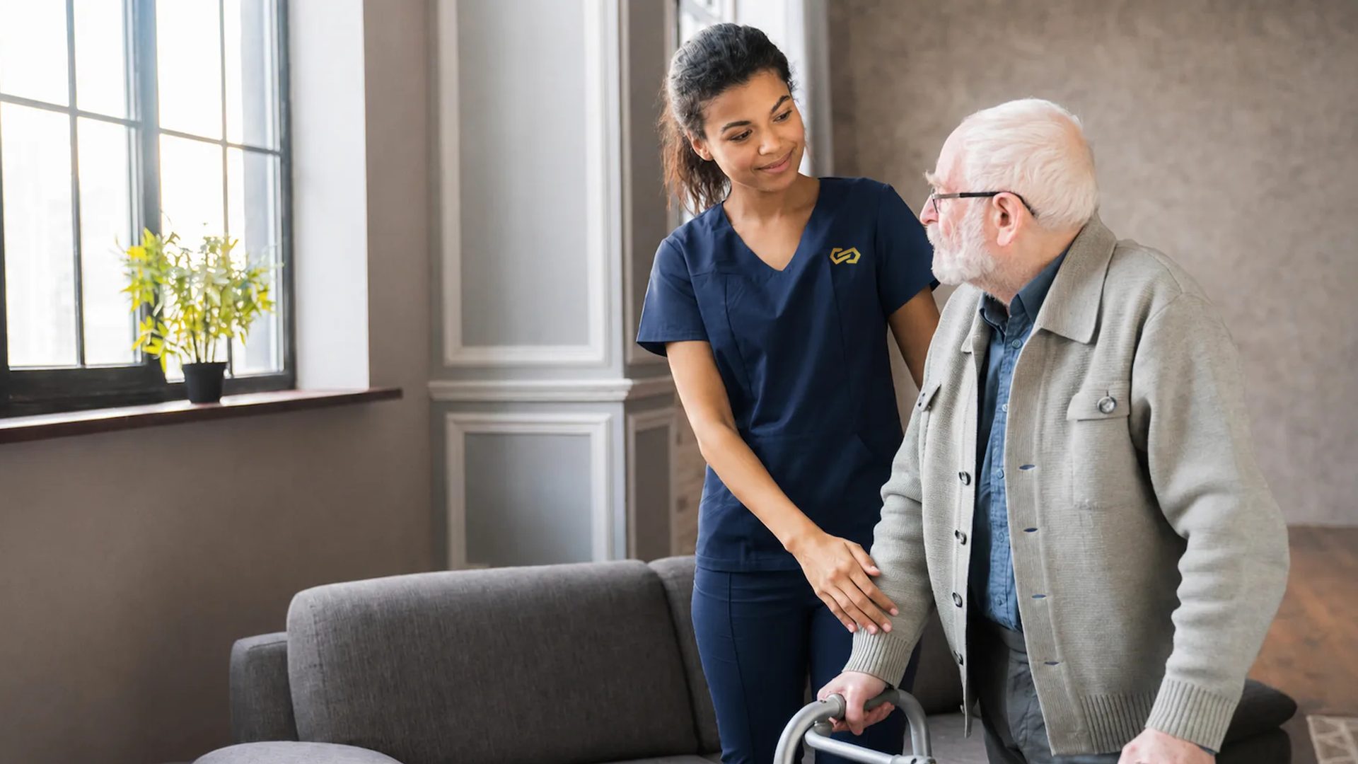 A nurse steadies an older man as he walks with a wheeled walking frame